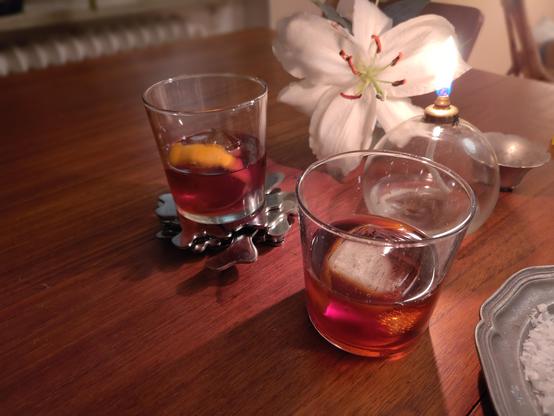 Två mörkröda drinkar med is och apelsinskal på ett teakbord. Bredvid en vit lilja och ett oljeljus.