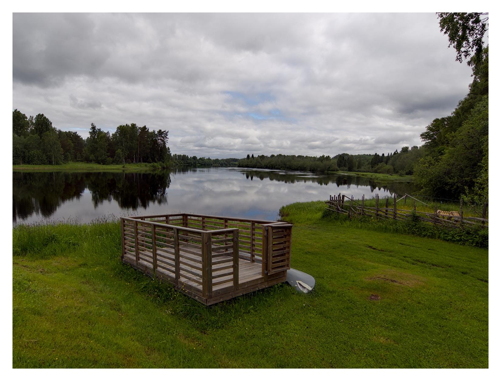 En liggande bild på en rektangulär träkonstruktion med en ingång, staket utan tak på en gräsmatta. Bredvid ligger en grå kanot med ett hajansikte målat på ena änden. I bakgrunden rinner Klarälven genom ett grönskande landskap. Himmelen är molnig med små, blå fläckar.
