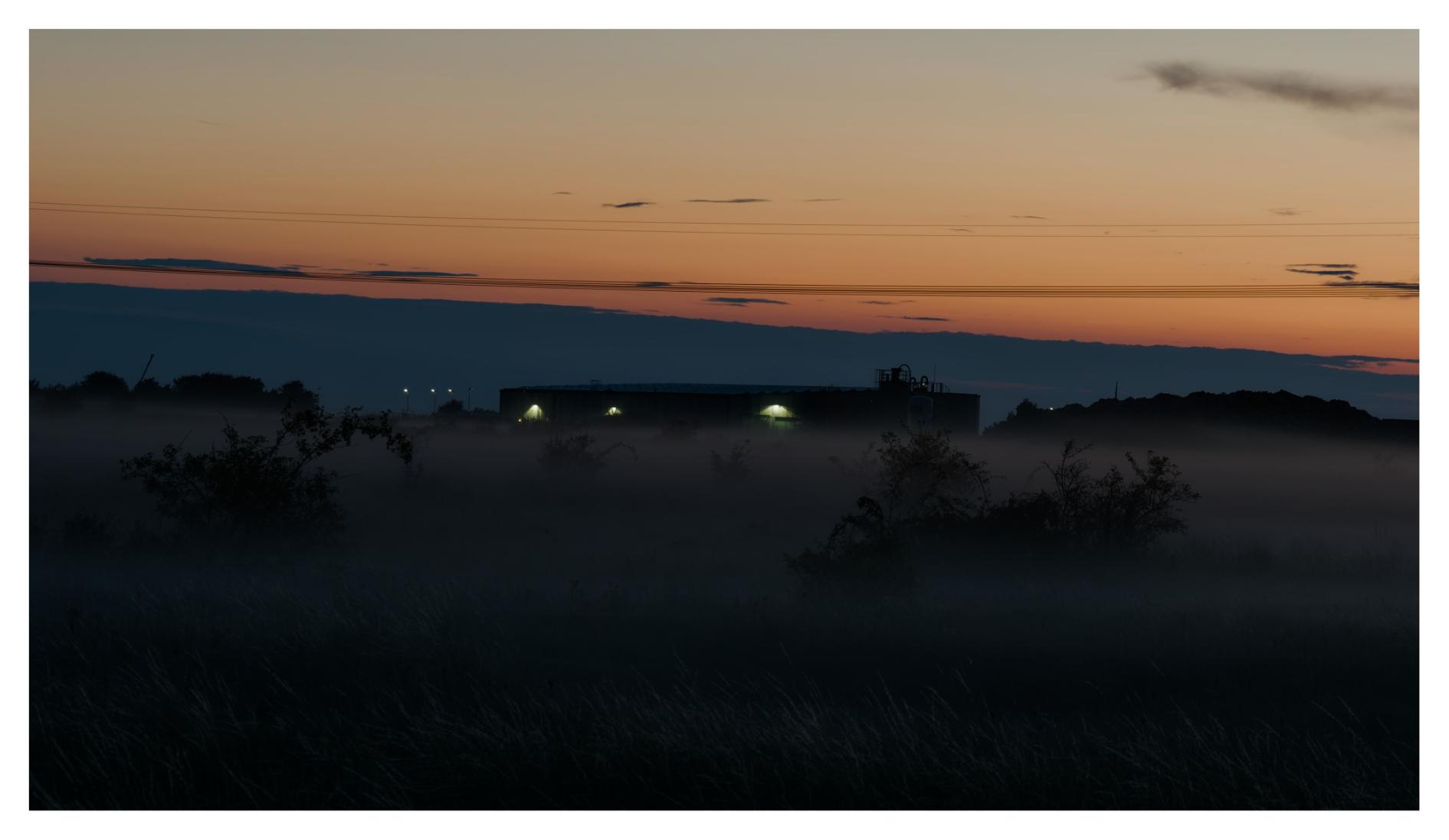 En liggande bild på en solnedgång över en äng. Längre bort står en byggnad med ljus tänt. Kablar går tvärs över himlen som har små moln.