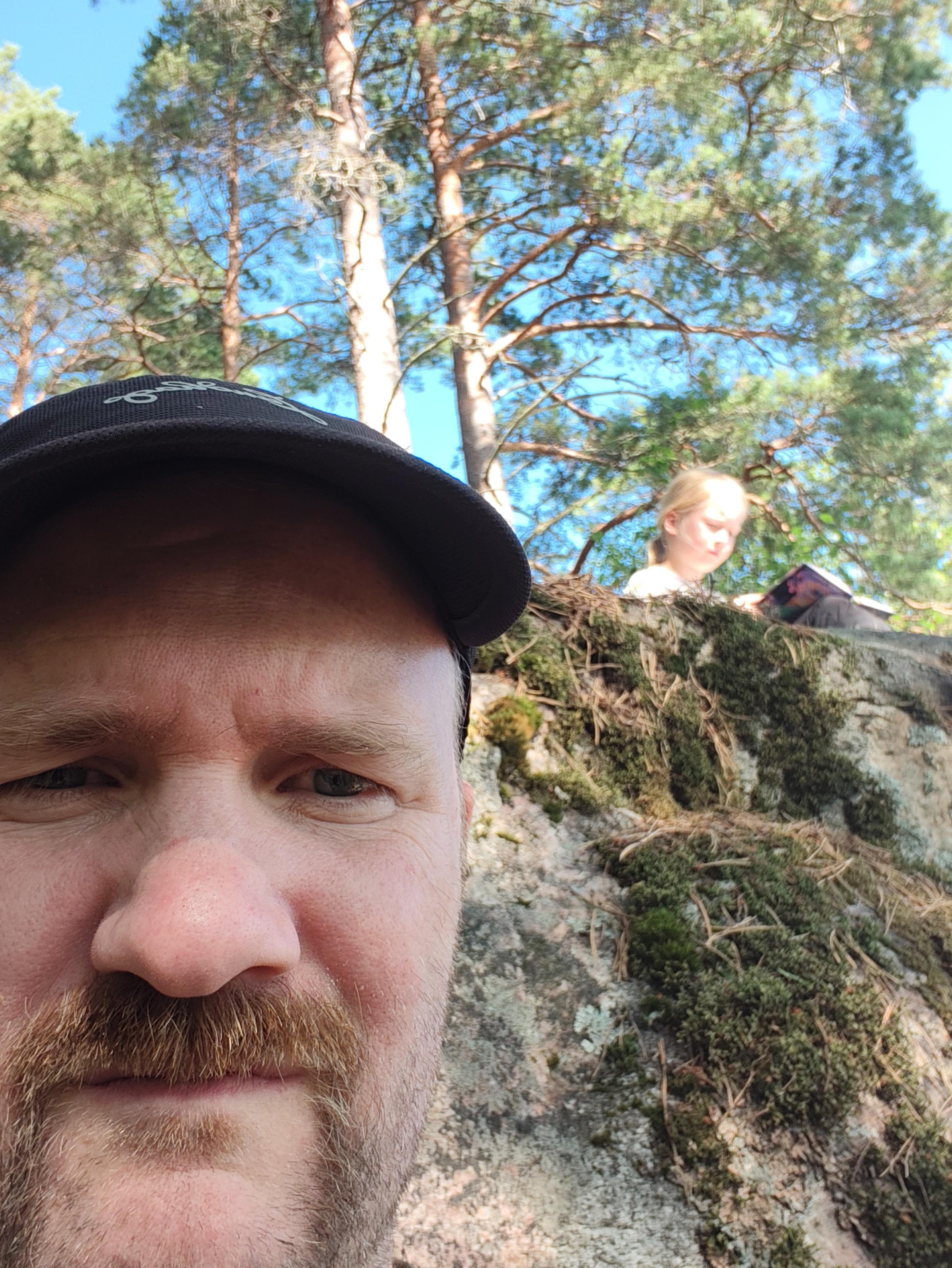 Selfie på en man i rockmustasch och svart keps. I bakgrunden är en sten varifrån det sticker upp ett antal tallar. Ett barn sitter på stenen och läser.