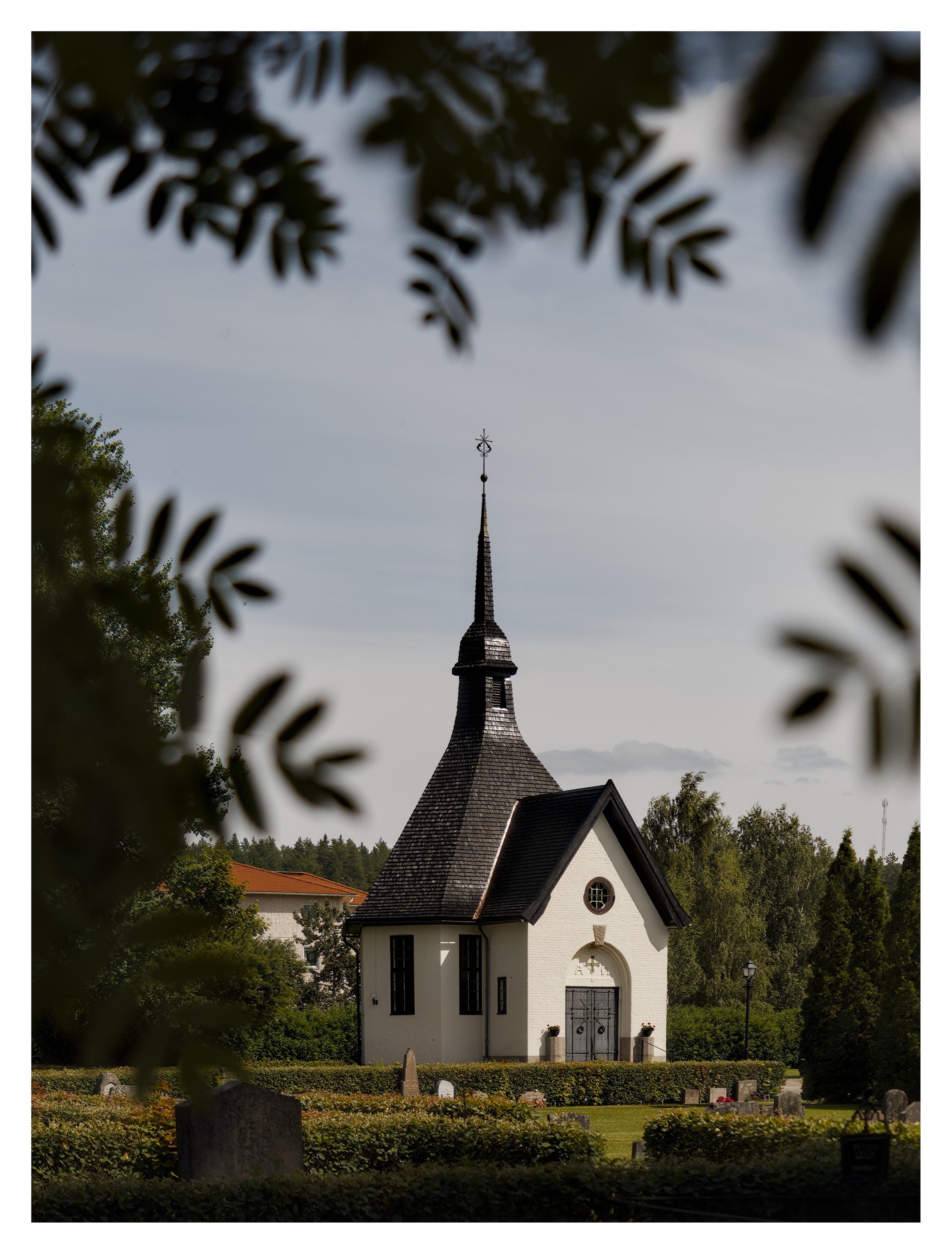En stående bild på ett vitt kapell med svart, spetsigt tak. Framför är många gravstenar på en kyrkogård. Bilden inramas av några grenar ur fokus i förgrunden.