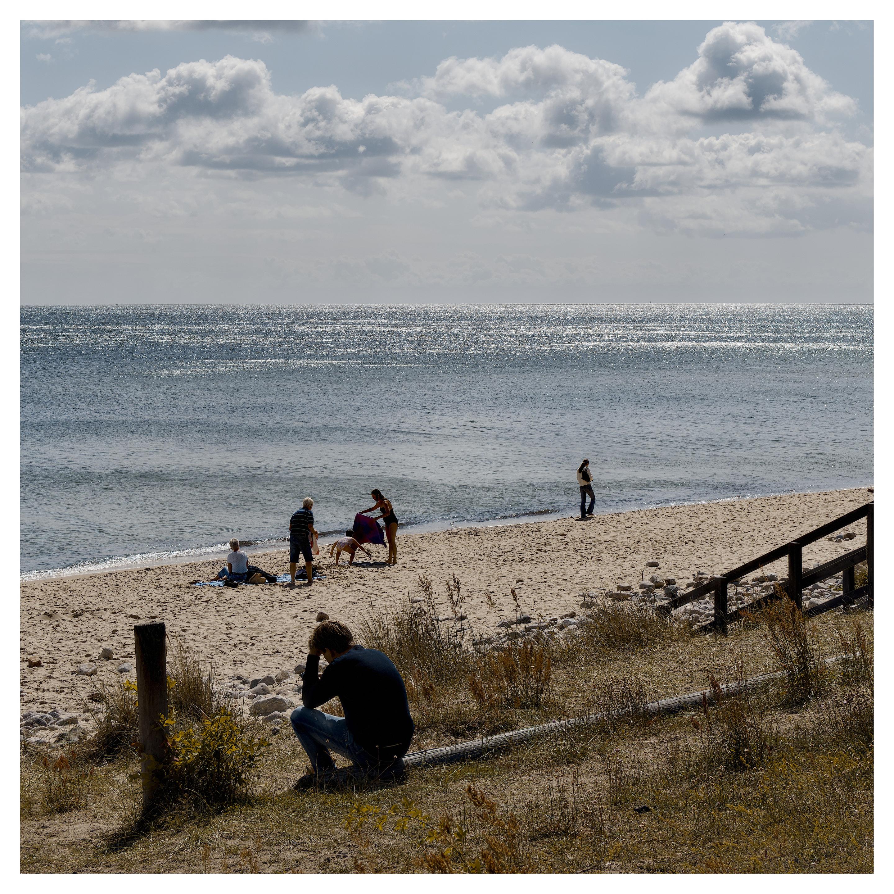 En fyrkantig bild på en sandstrand vid havet. Närmast sitter en ung man på en liggande stock med huvudet i en hand och tittar nedåt. Längre bort i sanden leker ett barn med sin familj och till höger går en ensam flicka med en väska över axeln. Himmelen har många stackmoln.