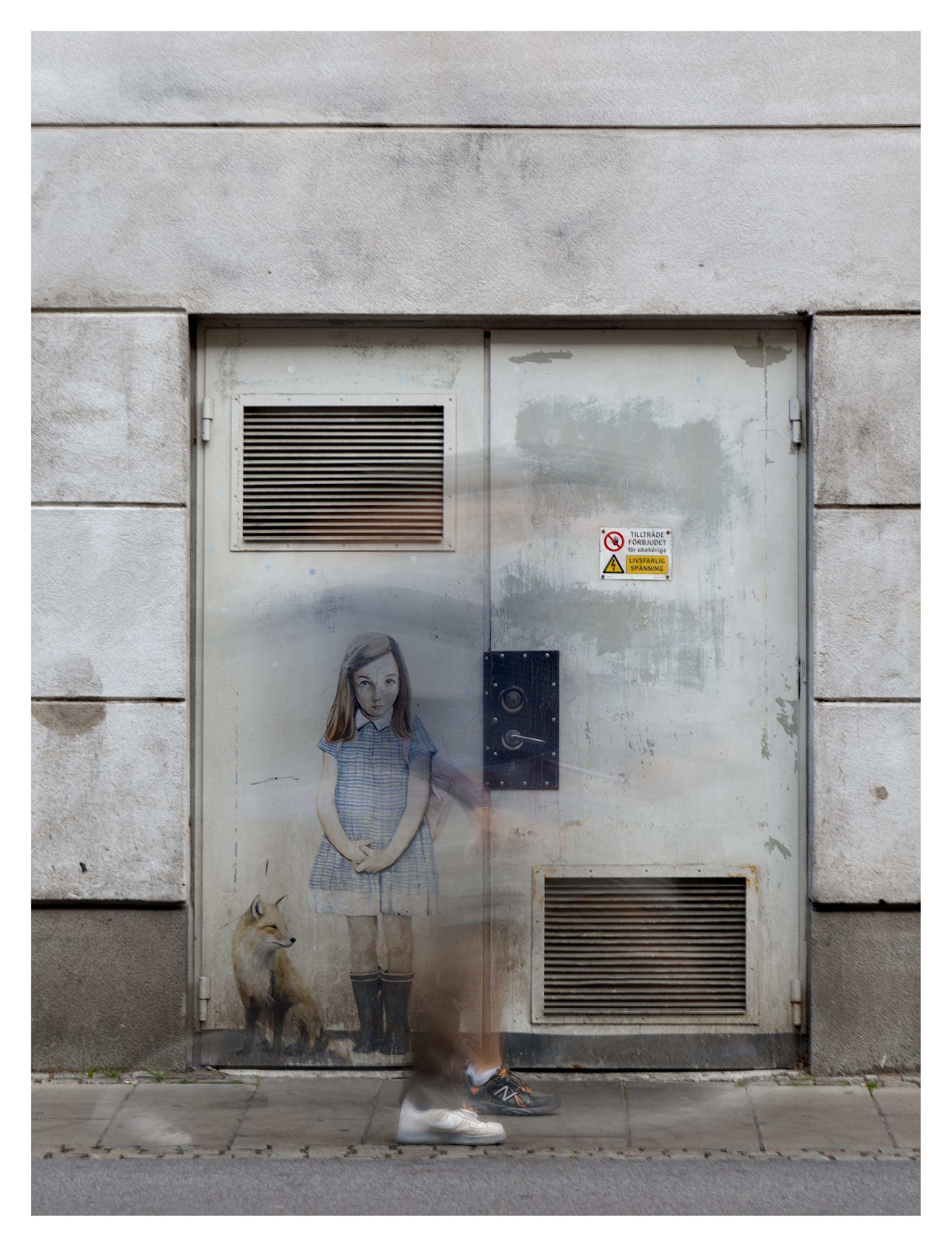 En stående bild på en vit dubbeldörrar med luftgaller. Runt handtaget är det svart. På vänstra dörren är en flicka i ljusblå klänning och mörkblå gummistövlar målad. Bredvid sig har hon räv. Framför dörren passerar två människor, men eftersom bilden är tagen med lång slutartid är endast skorna någorlunda skarpa.