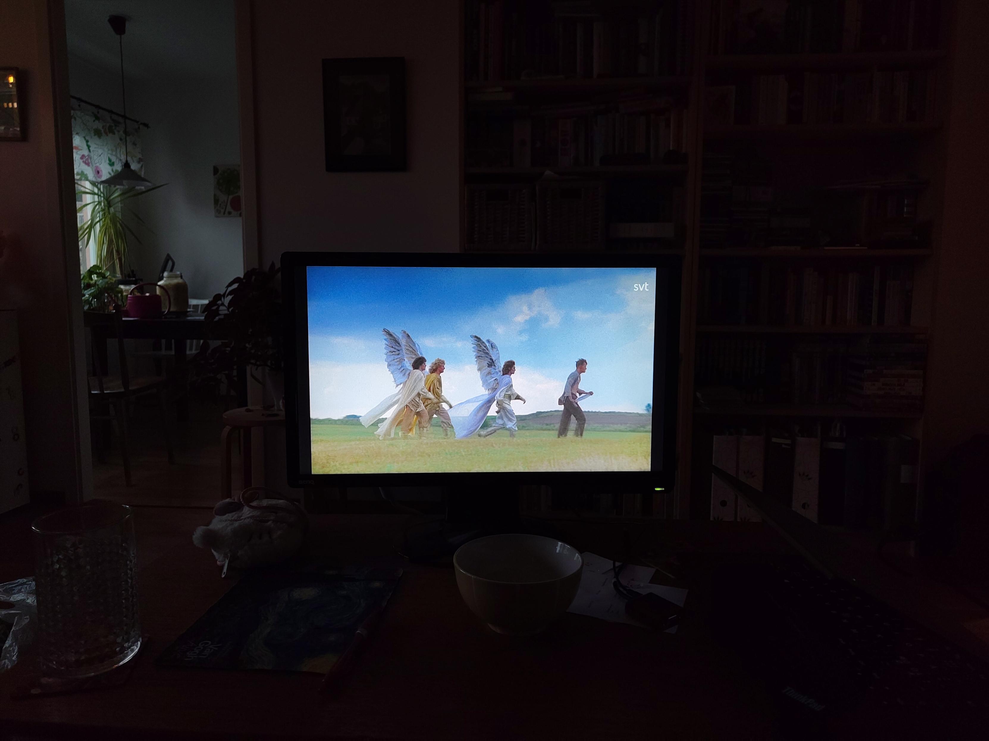 Ett mörkt rum med en skärm stående på ett bord. Skärmen visar en av de mer ikoniska scenerna från filmen Den enfaldige mördaren där huvudkaraktären Sven går över ett färt med en stor kniv följt av tre änglar.