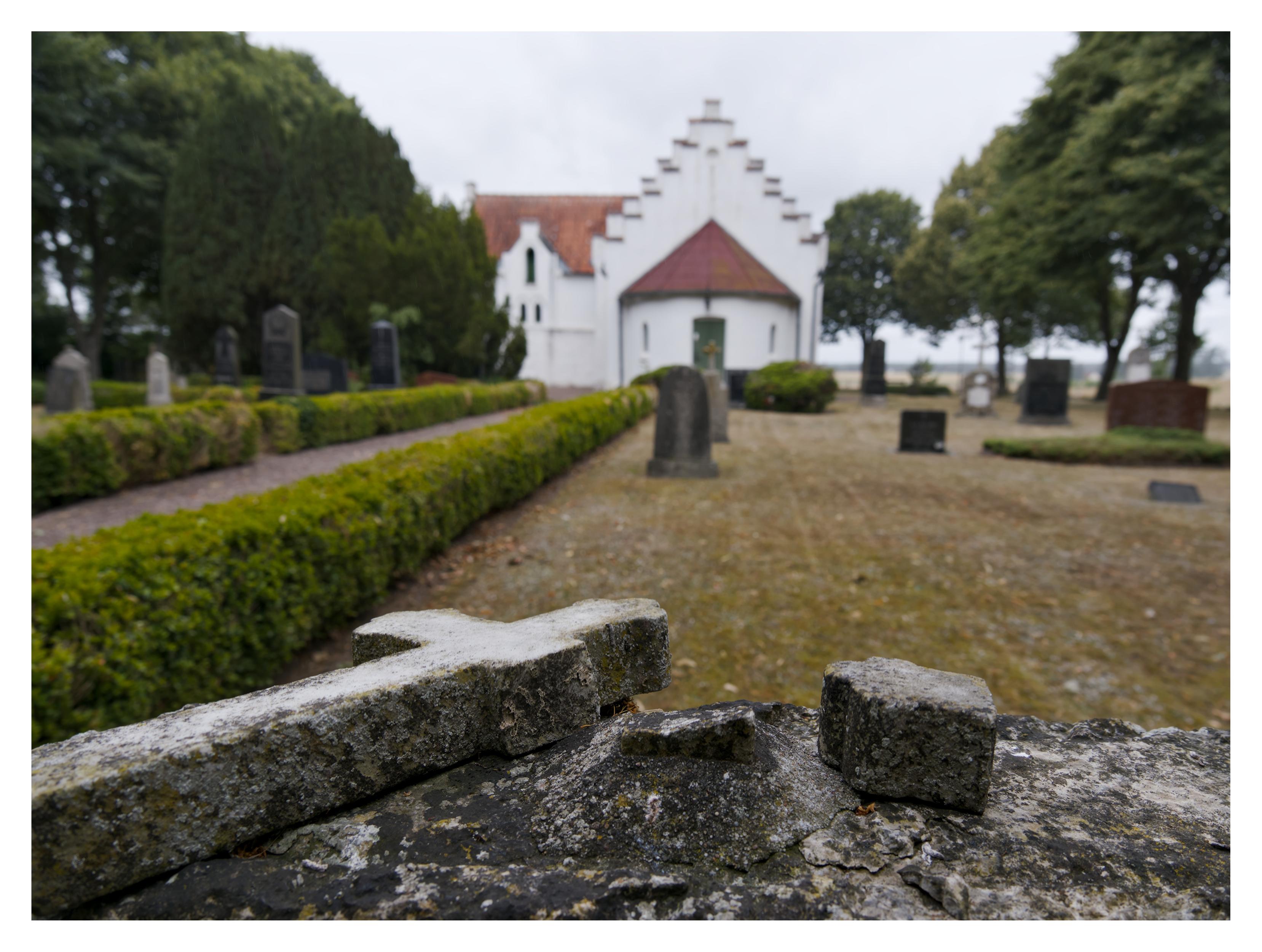 En liggande bild på en vit stenkyrka med rött tegeltak. I förgrunden att avbrutet kors som ligger ovanpå sin gravsten. Till vänster går en grusad gångväg mellan två gröna häckar. Fram till kyrkan står gravstenar glest på en gräsmatta. Längs sidorna på bilden finns gröna lövträd. Himmelen är molnig.