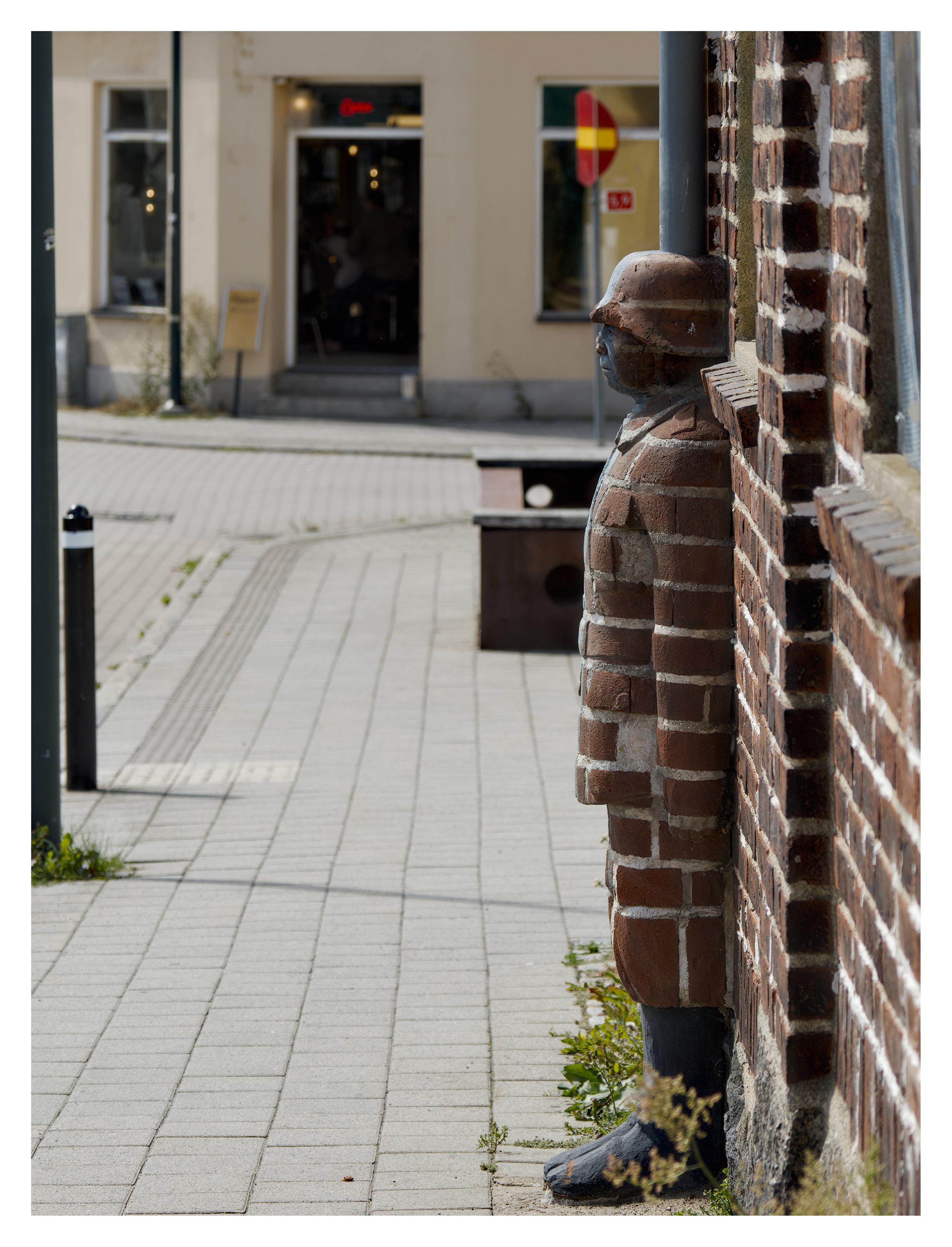 En stående bild på en figur av en soldat som står mot en husvägg i tegel på höger sida. Soldaten är i samma tegel. Bilden är tagen från sidan. På vänster sida går en trottoar och på andra sidan gatan rakt fram är en affär och en röd gatuskylt med ett gult streck.