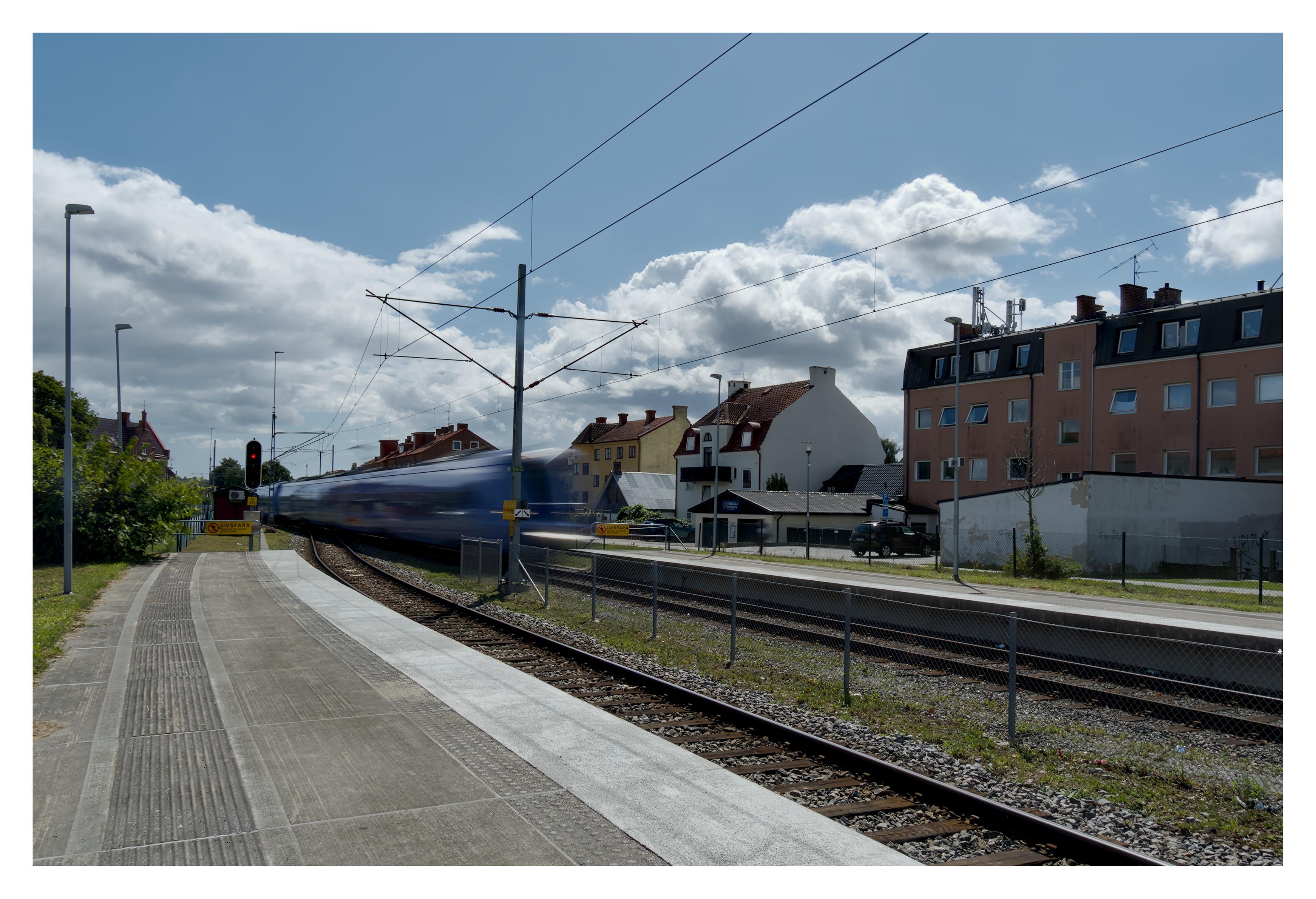 En liggande bild på ett suddigt, lila tåg kommer farande från vänster vid en plattform med knottrig och räfflad yta.Till vänster en signal med rött ljus. Till höger byggnader och en parkerad, svart bil.