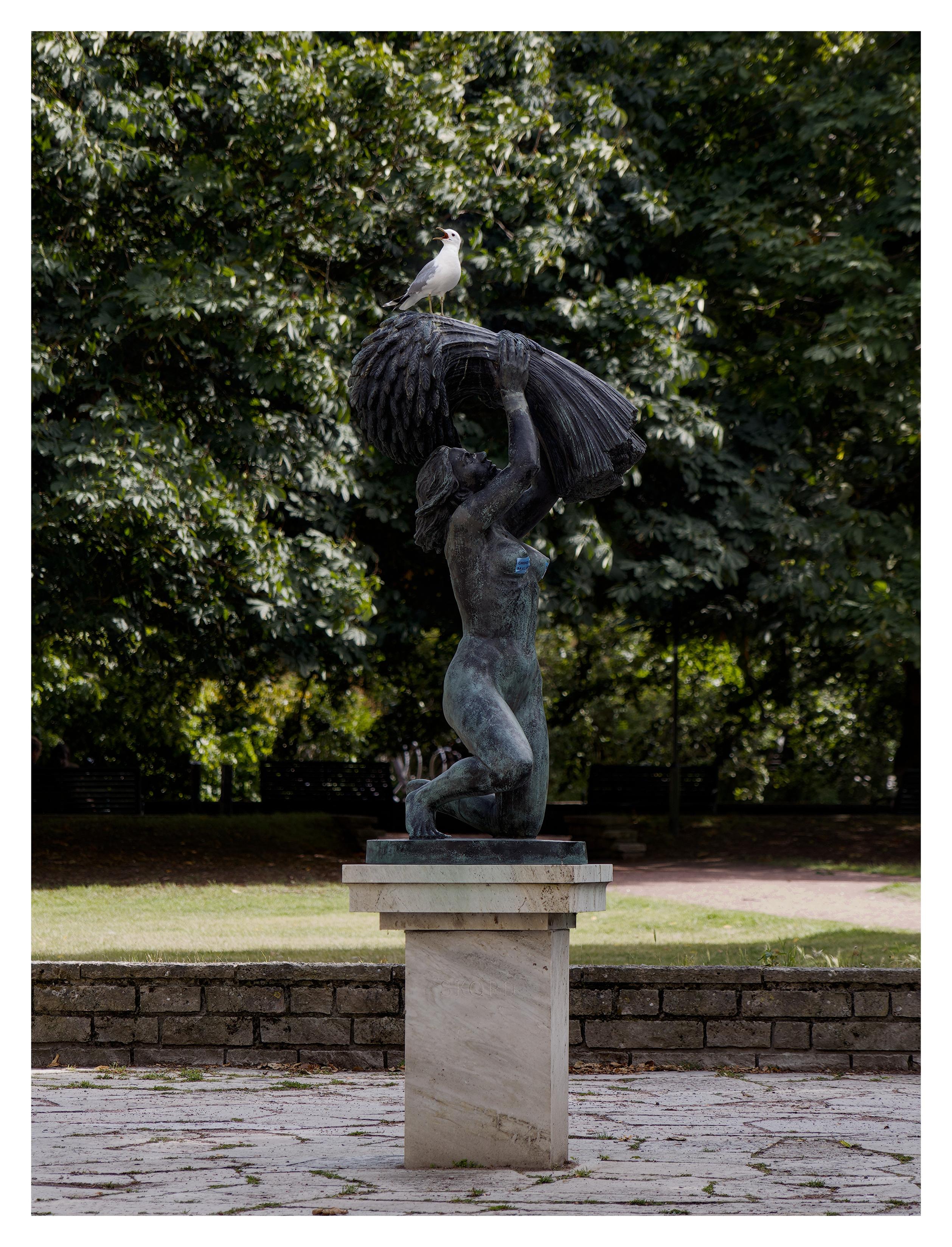 En stående bild på en bronsstaty på en sockel av sten. Statyn föreställer en naken kvinna på knä som håller upp en kärve. På toppen sitter en levande fiskmås och skriker. En park med träd i bakgrunden.