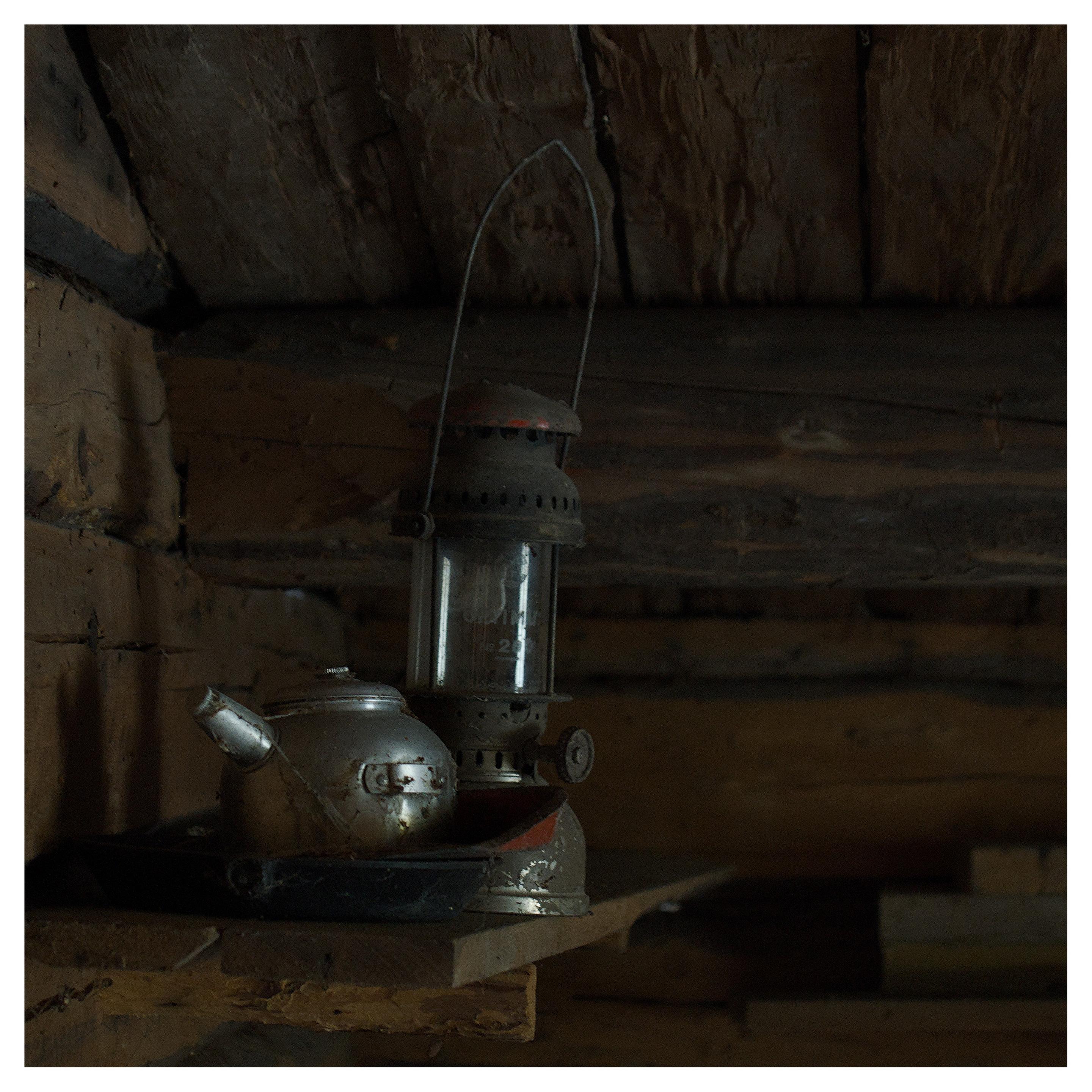 En fyrkantig, lite mörk bild på en gammal kaffekanna i metall och en fotogenlampa. De står på en trähylla i ett trähus.