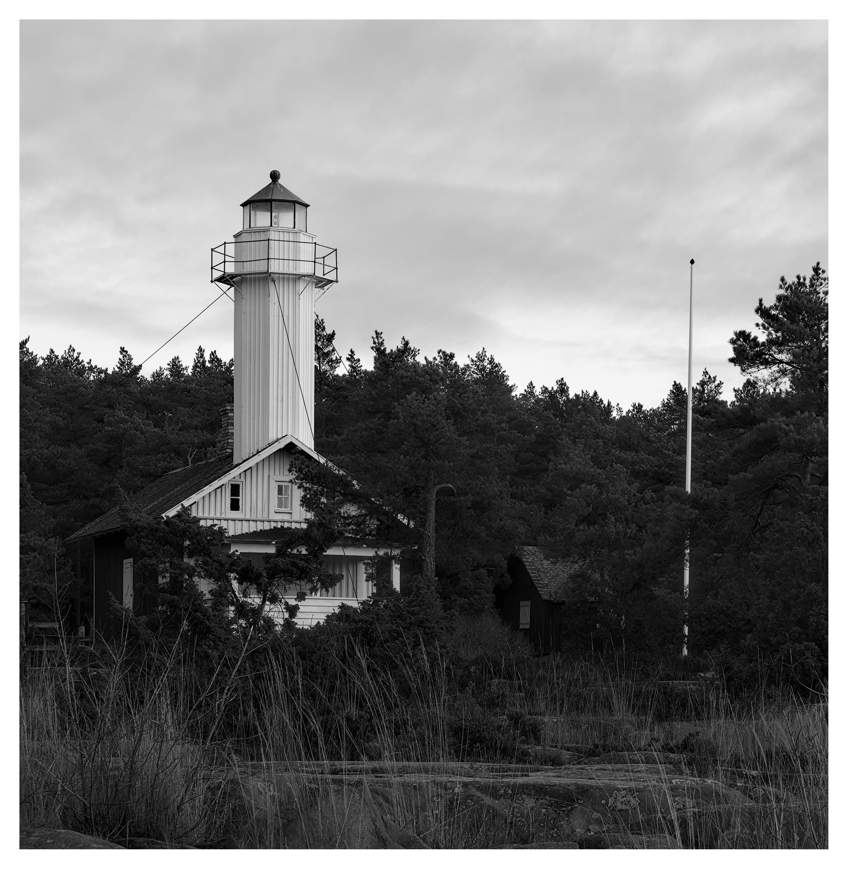 En svartvit fyrkantig bild på en fyr som står i en bebott hus. Till höger en ekonomibyggnad och en flaggstång. I förgrunden glest, högt gräs vid stenhällar. I bakgrunden barrskog. Himmelen är molnig.