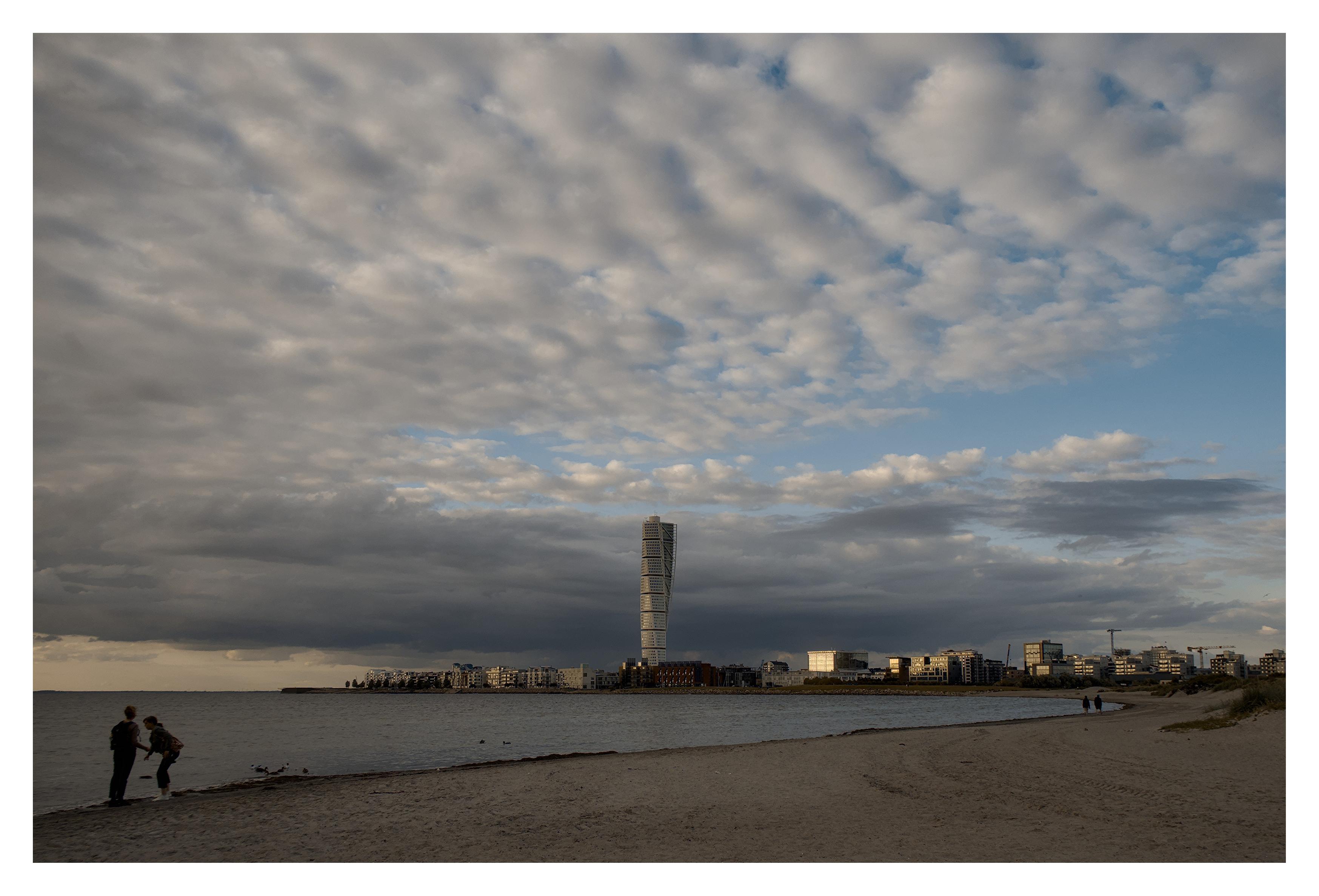 En liggande bild på det skruvade huset Turning Torso i Malmö. Sandstrand med några människor i förgrunden och molnig himmel med blå luckor här och var. Kvällssolen skiner och blänker i alla hus andra sidan viken.