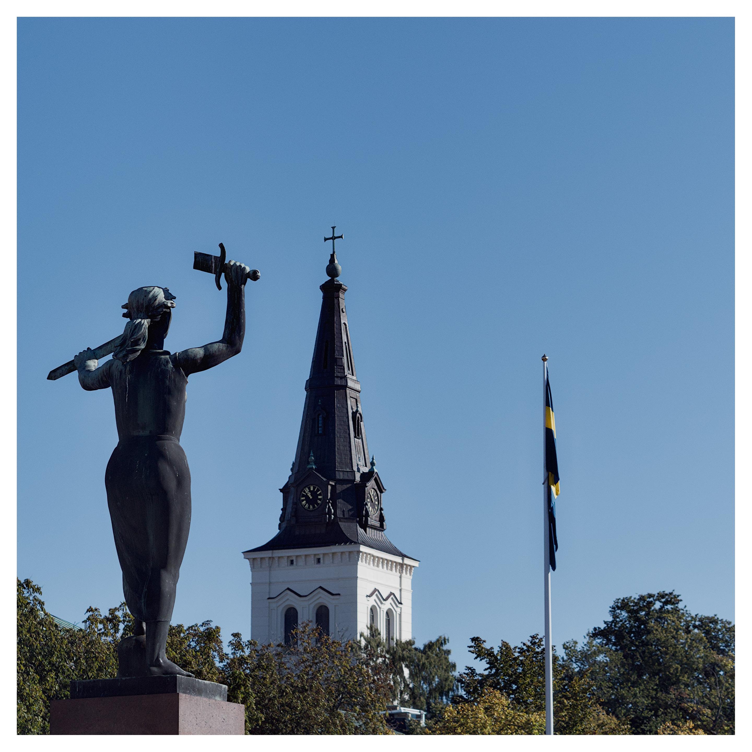 En fyrkantig bild på en staty, ett kyrktorn och en slak sverigeflagga på en vit flaggstång. Statyn är en kvinna som håller ett avbrutet svärd i sina händer. Undre delen av bilden har topparna av lövträd. Himmelen är klarblå och molnfri.