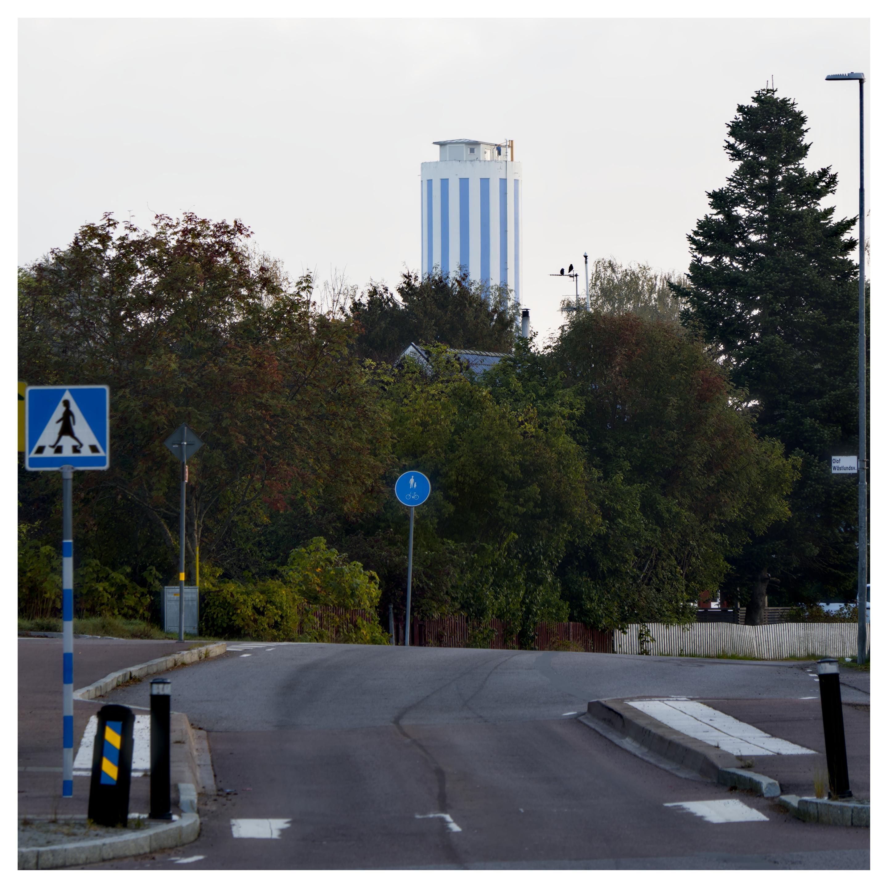 En fyrkantig bild på ett vattentorn med vertikala ränder, ljusblått och vitt. I förgrunden ett övergångsställe med en bå skylt, längre fram på gatan en blå skylt för gång- och cykelväg. Sedan träd fram till vattentornet. Himmelen jämngrå.