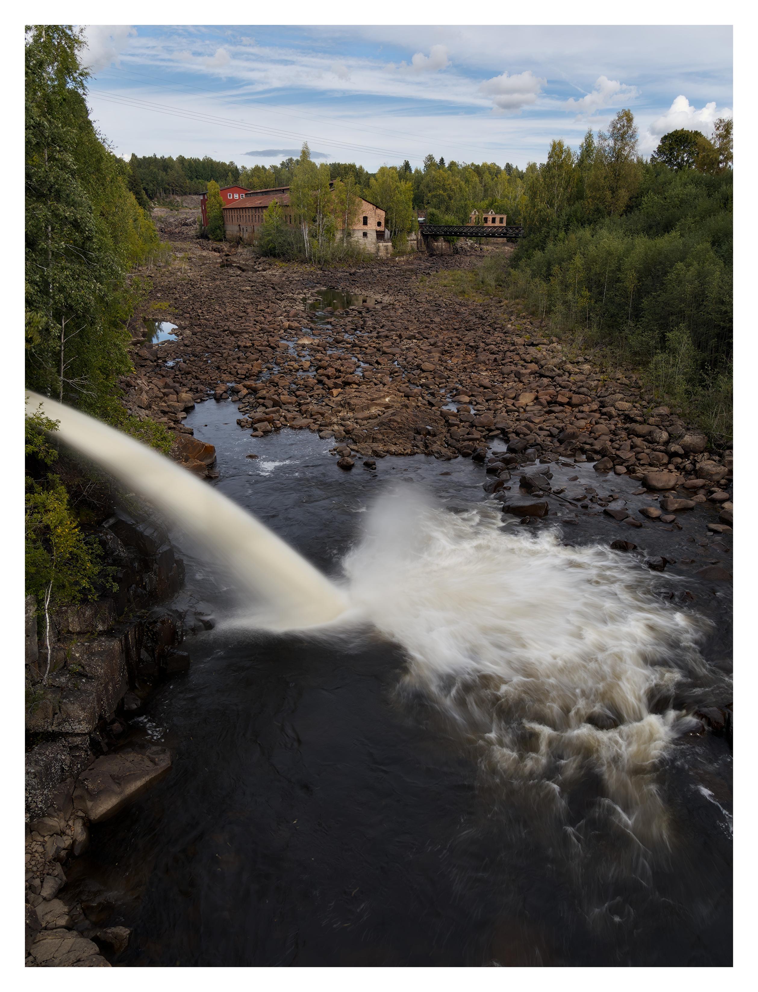 En stående bild på vatten som forsar ner från vänster i en bassäng vatten. Älven ovanför är torrlagd och består bara av sten. Längre upp är några byggnader där älven delar sig i två. I den högra grenen är en bro. Skog på båda sidor älven, himmelen är nästan fylld med vita slöjmoln.