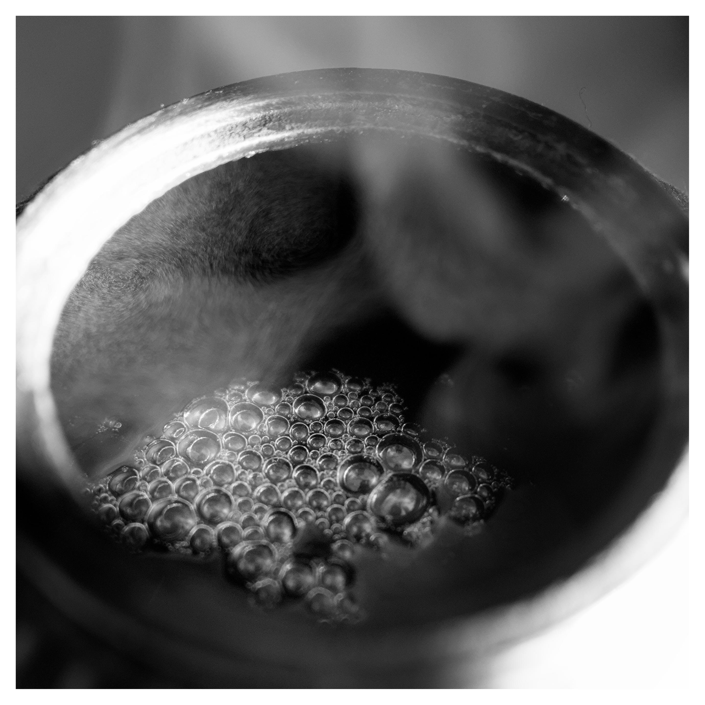 En fyrkantig, svartvit bild på en tekanna snett uppifrån. Rök från det heta vattnet och många bubblor på vattnet.