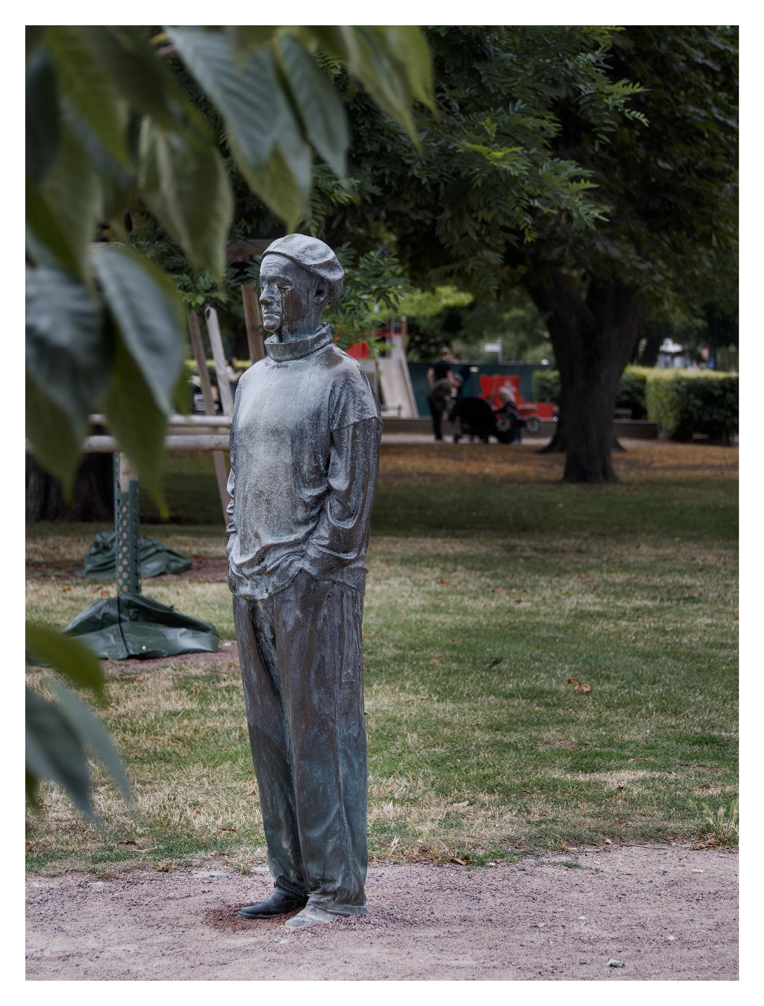 En stående bild på en staty av en man med en keps på huvudet och händerna i byxfickorna. Han står i sand i en park. Blad hänger ner från vänster i förgrunden, några träd i bakgrunden.