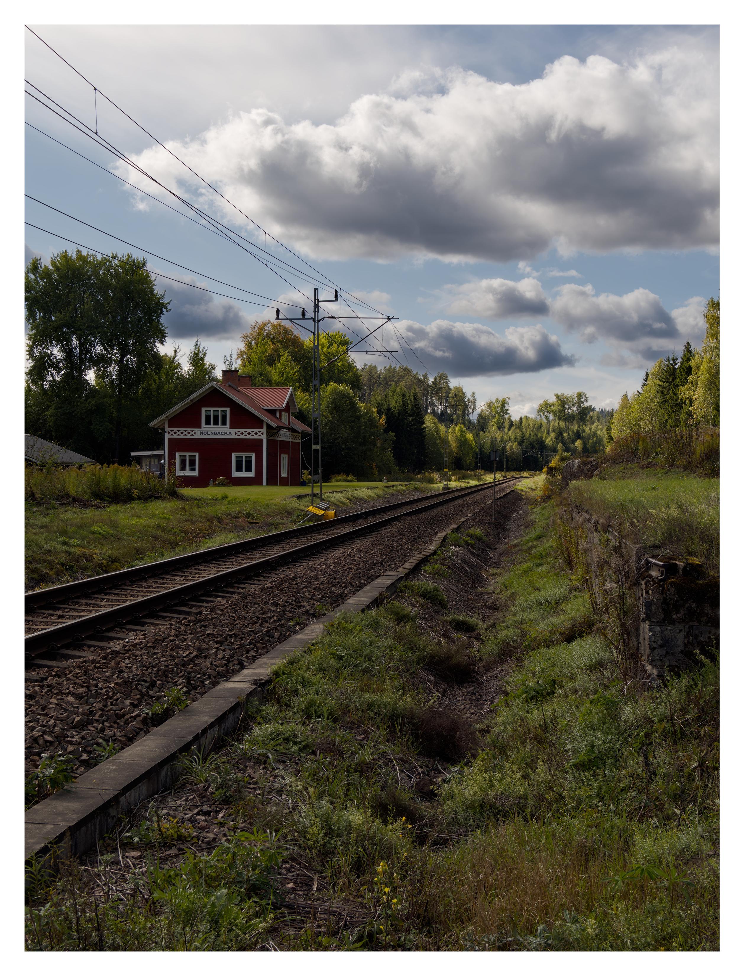 En stående bild på järnvägsspår som kommer in från nere till vänster och försvinner i fjärran. På vänster sida ett rött stationshus i trä med vita knutar och texten "MÖLNBACKA" på väggen. Till höger en överväxt liten betongmur. Till vänster om spåret ligger en gul skylt med texten "Smörjapparat". Ganska mycket moln på en annars blå himmel.