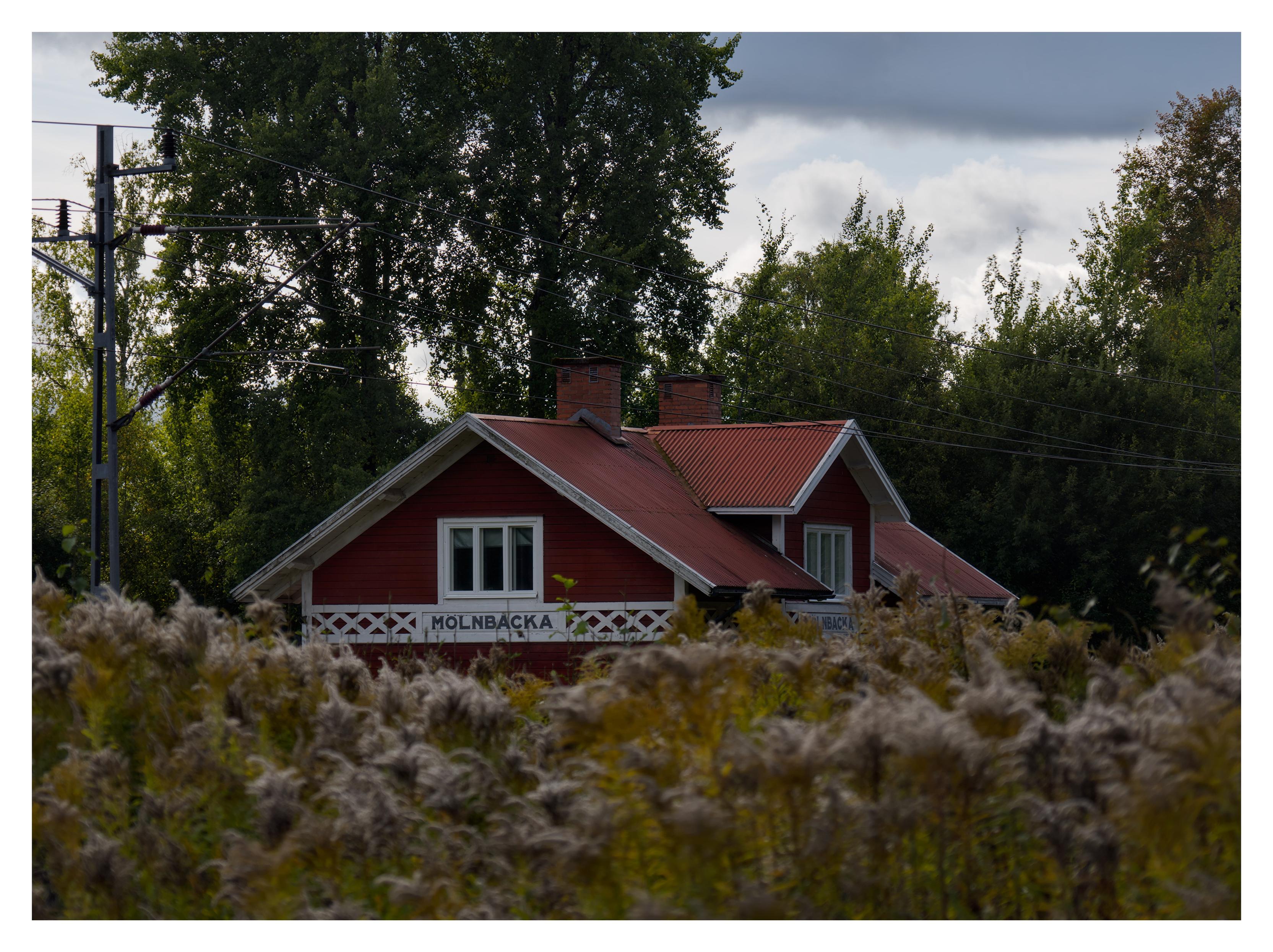 En liggande bild på överdelen av stationshuset i rött trä med vita knutar. Här visas texten "MÖLNBACKA" både på kortsidan och framsidan. Växter ur fokus i förgrunden, träd i bakgrunden och järnvägens kablar i luften. Mestadels moln på himmelen.