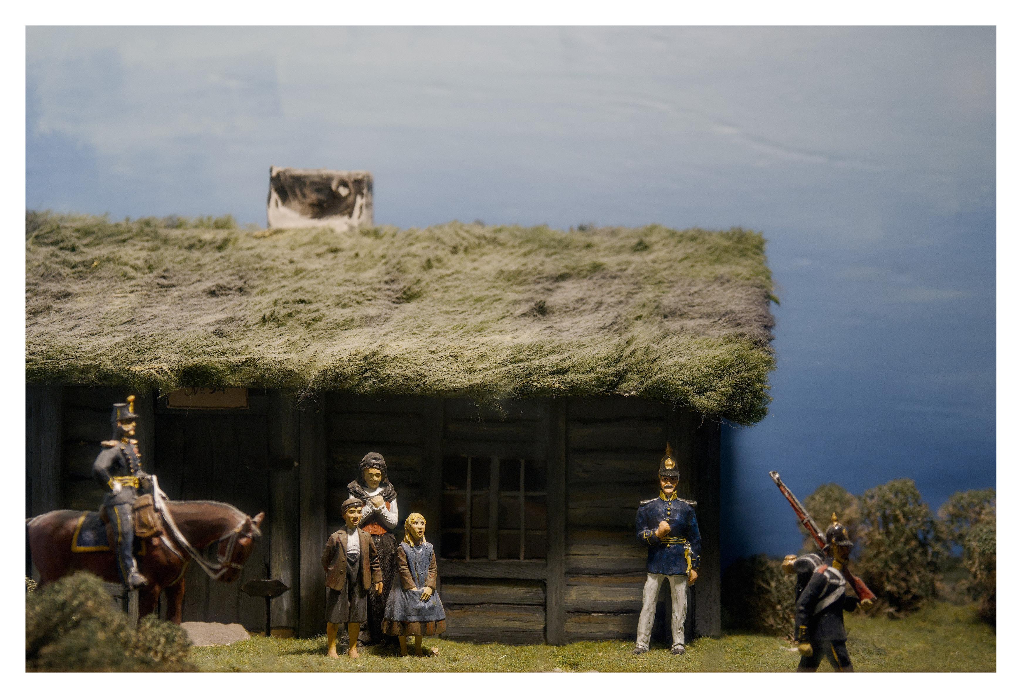En liggande bild på en modell av en trästuga med gräs på taket och soldater utanför. Em mor med två barn står utanför huset. Det ska förmodligen föreställa 1800-talet.